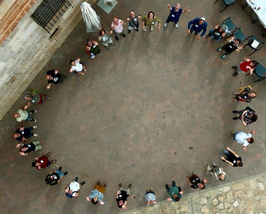 Bienvenida_Encuentro los asistentes al evento colocados en círculo en la plaza de Luco saludan sonrientes a la cámara a 10m por encima de sus cabezas.